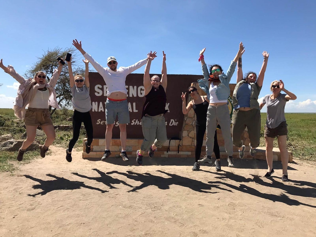 A group of individuals is captured mid-jump in front of a sign marking the entrance to a national park. Their joyful movements create elongated shadows on the ground, with a vast grassy landscape visible in the background.