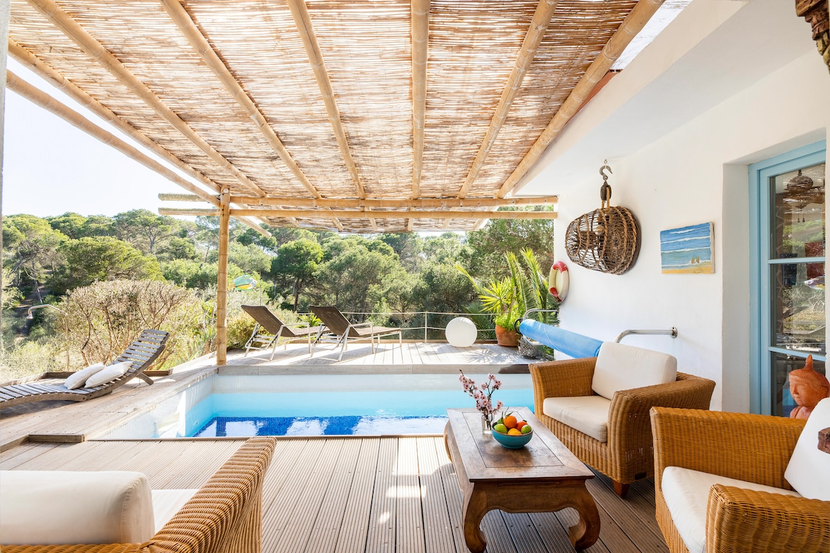 A spacious terrace is equipped with comfortable seating and a wooden coffee table, featuring fruit. A small pool reflects the sunlight, surrounded by lounge chairs. Lush greenery is visible in the background, adding to the serene environment.