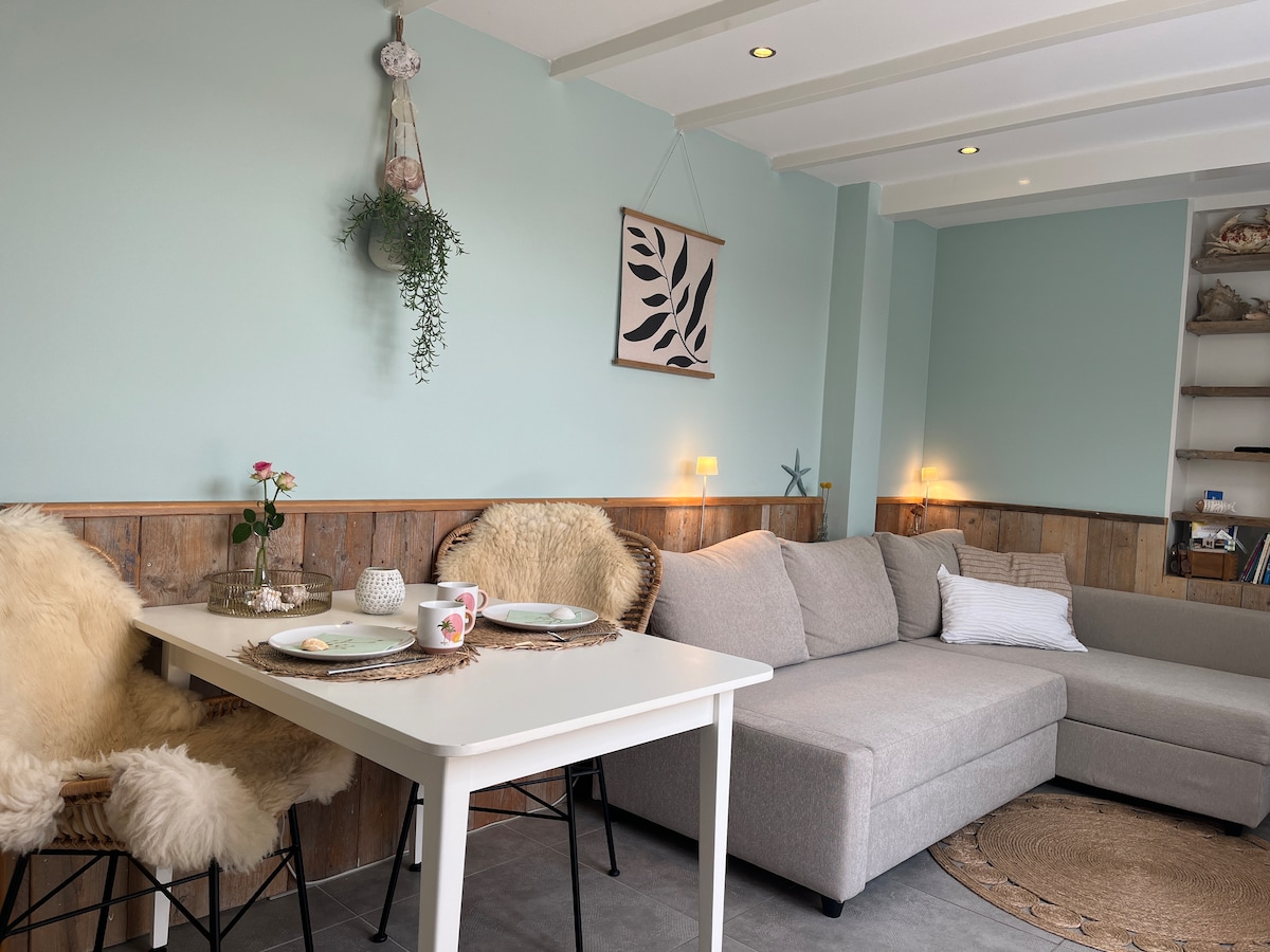 A cozy living area features a light green accent wall, a comfortable gray corner sofa, and a dining table set for two. Soft fur-covered chairs add warmth, while gentle lighting creates a relaxed ambiance. Decorative elements include wall art and a hanging plant.