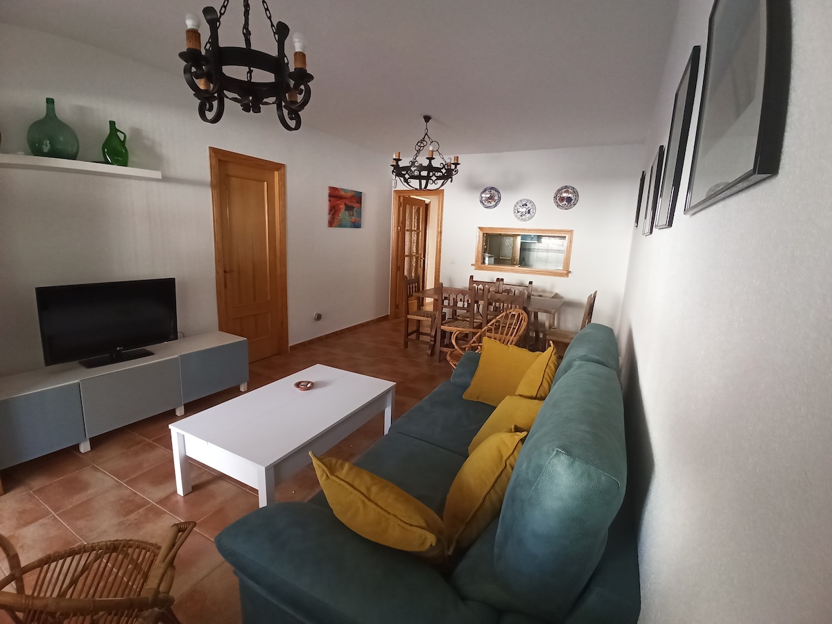 A living room area features a turquoise sofa adorned with yellow cushions. A coffee table sits in front of the sofa, while a television is positioned on a light-colored media unit. Light wood accents are present in the dining area, which includes a wooden table and chairs.
