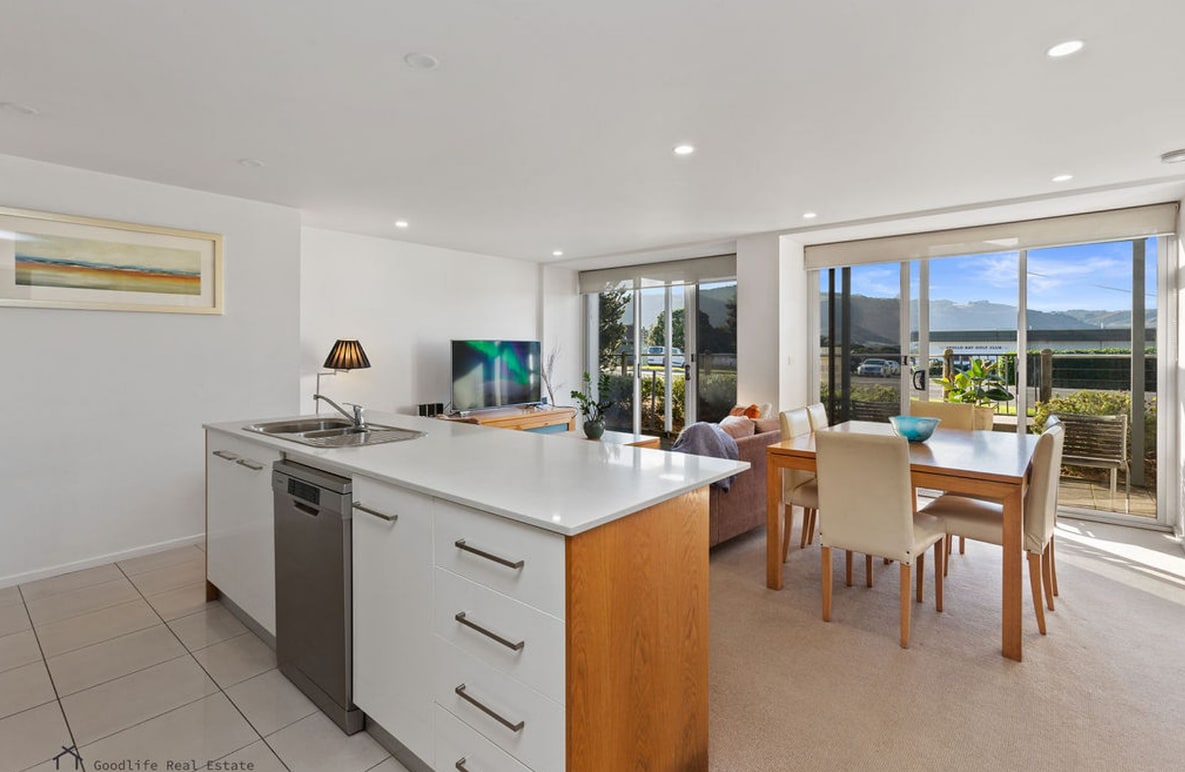 A modern kitchen is presented with a sleek island counter and stainless-steel appliances. Across, a dining area features a wooden table surrounded by light-colored chairs. Large glass doors enable bright natural light and provide views of outdoor seating and greenery.