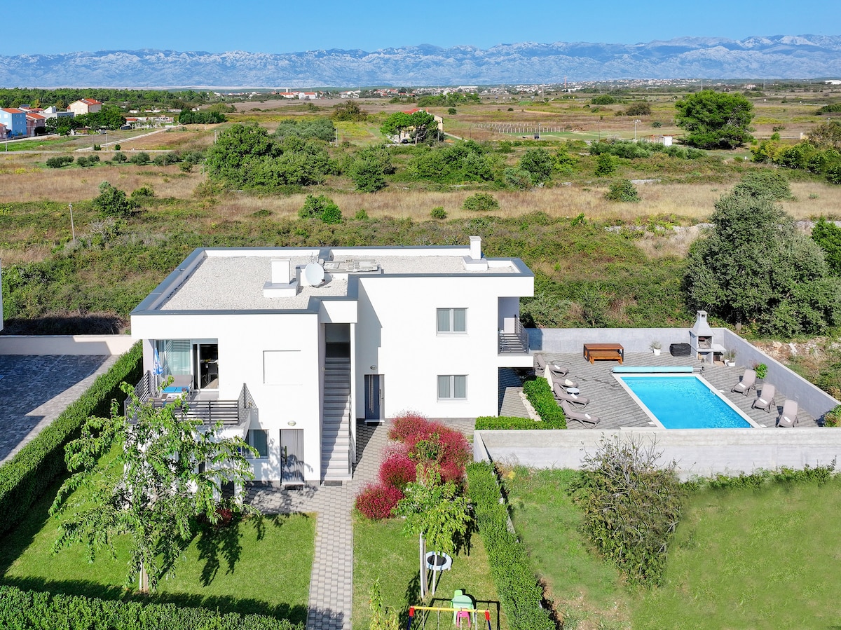 The villa is presented from an elevated perspective, showcasing its modern architecture and surrounding greenery. A swimming pool is visible, along with patio seating. The garden features vibrant landscaping, and a playground is seen in the corner, providing a family-friendly environment.