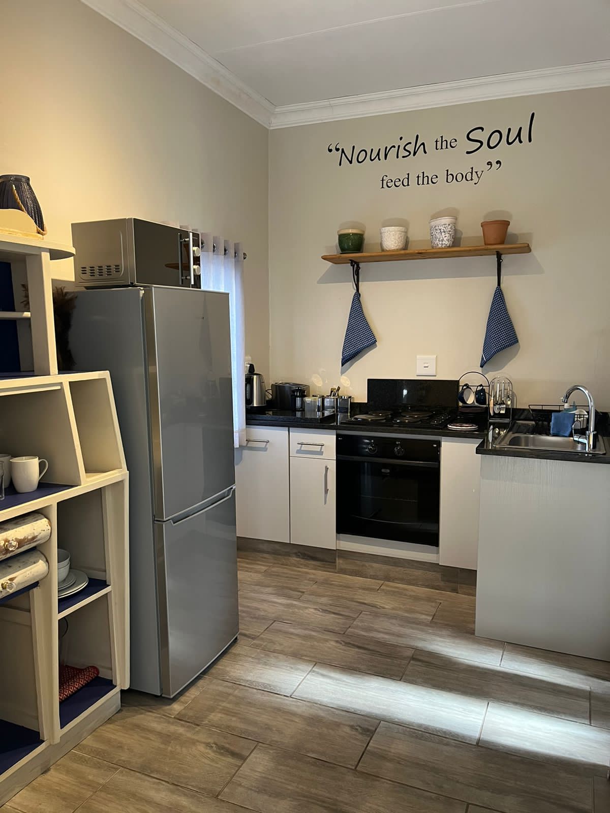 A fully equipped kitchen features modern appliances, including a refrigerator and stove. Shelves display various mugs and decorative items, while sleek countertops provide ample space for meal preparation. Light-colored cabinetry enhances the bright and functional atmosphere.