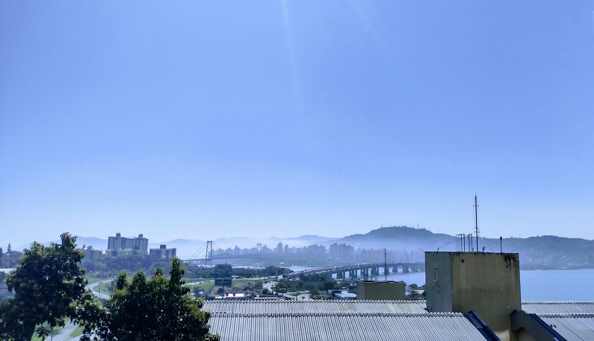 A panoramic view of the city skyline is captured under a clear blue sky. The distant mountains provide a subtle backdrop, while a bridge spans across the water in the foreground. Lush greenery is visible in the lower part of the image.