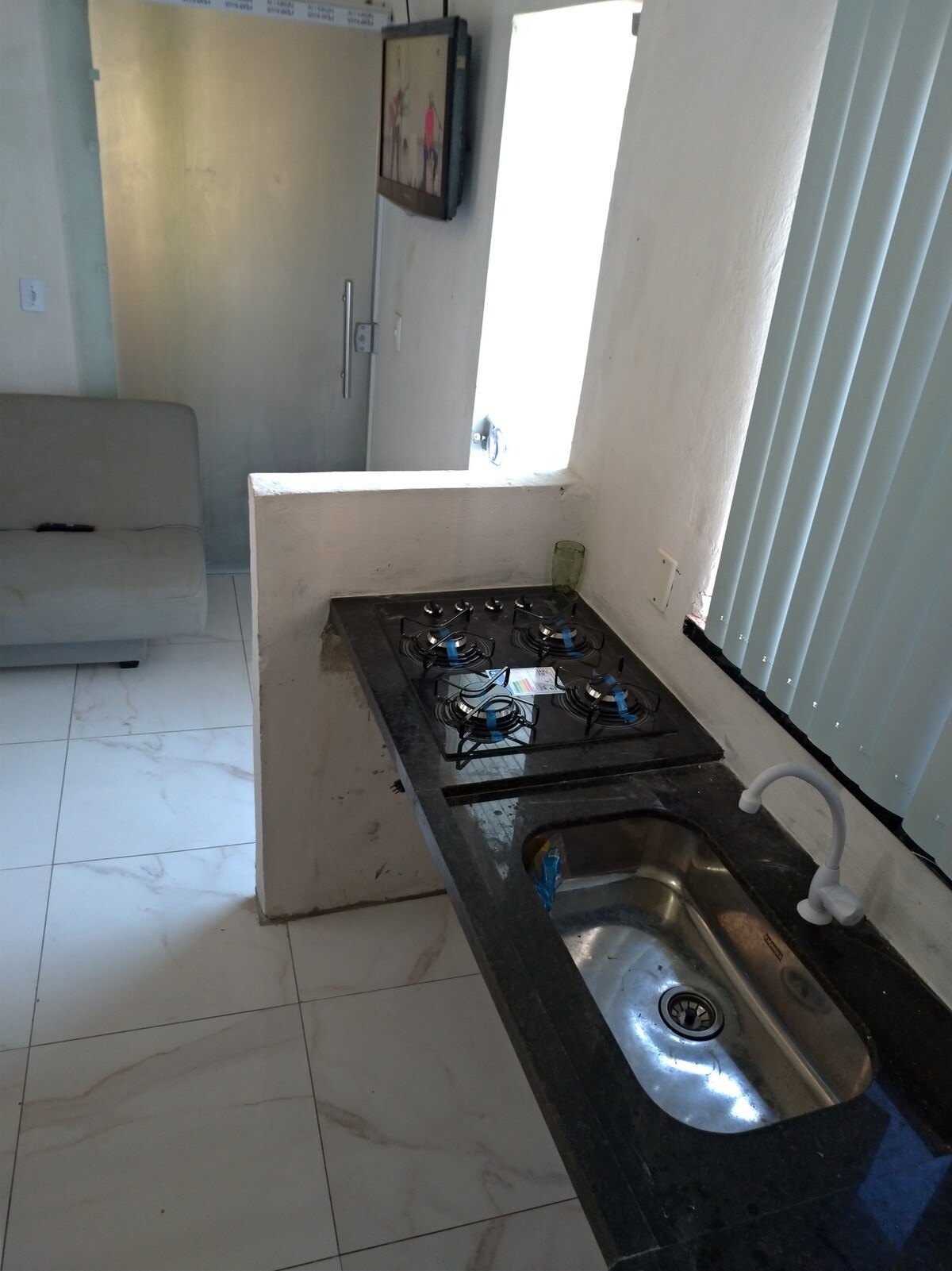 A functional kitchen area is depicted, featuring a countertop with a sink and a four-burner gas stove. Natural light is allowed in through a nearby window, and a minimalist sofa is positioned in the background, enhancing the kitchen's practicality.
