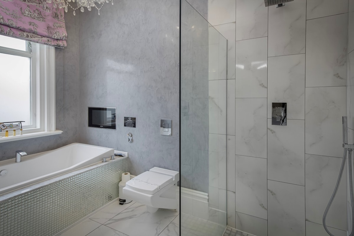 The luxurious bathroom features a two-person spa bath, equipped with a built-in TV. Elegant marble tiles adorn the space, complemented by a glass shower enclosure. A stylish chandelier hangs above, while a window offers natural light, framed by patterned curtains.