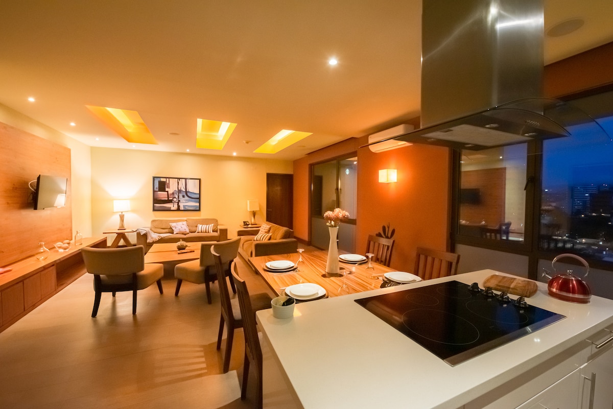 The open-concept living area features a modern kitchen with a sleek countertop and induction cooktop. A dining table is set for four, while the cozy seating area includes a sofa and armchairs, all illuminated by soft lighting that enhances the warm atmosphere.