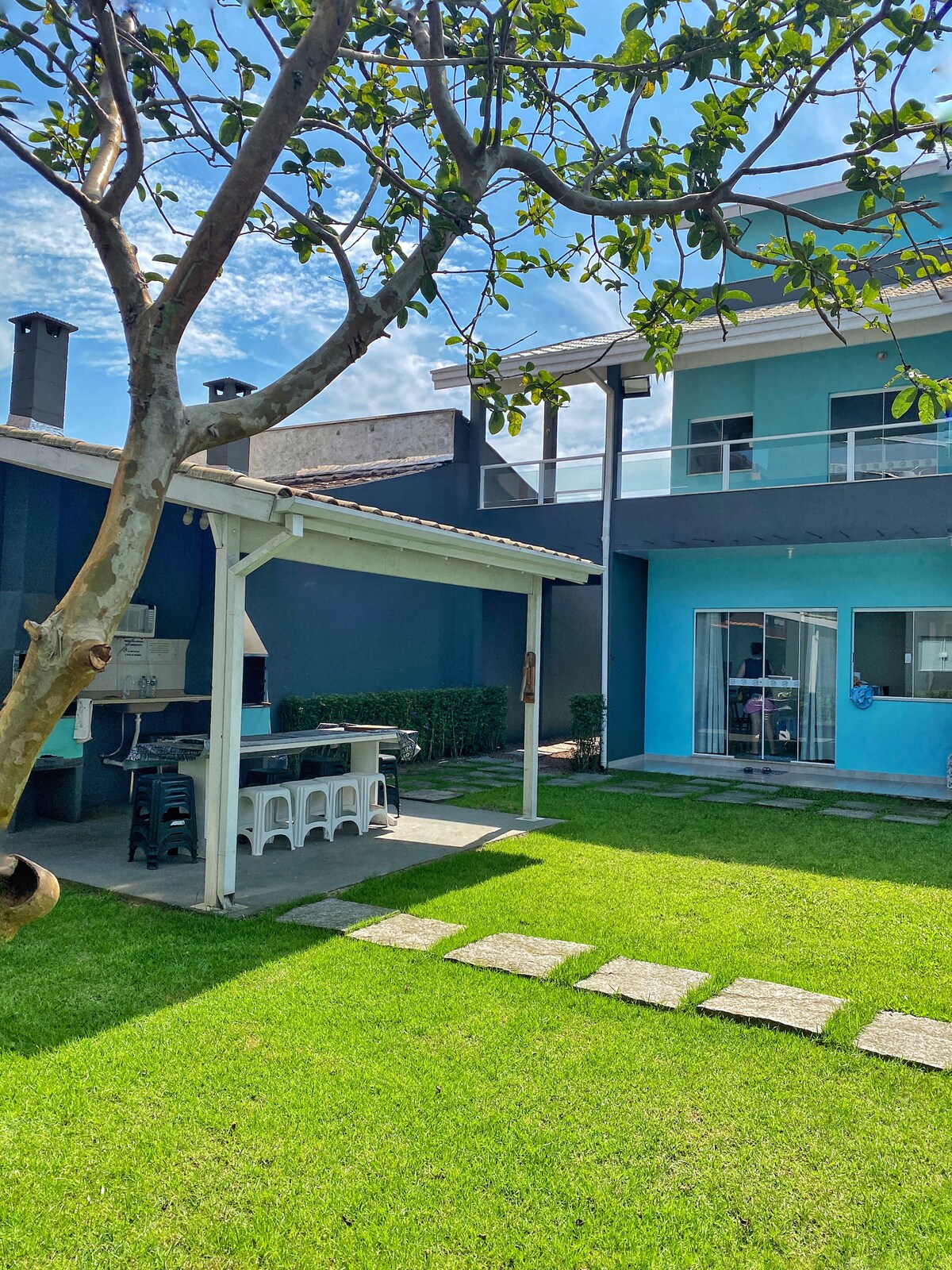 A spacious outdoor area features a well-maintained lawn with stone pathways leading to a covered barbecue area. The structure is painted in light blue, with large glass doors that provide a view into the interior space, and a tree provides gentle shade.