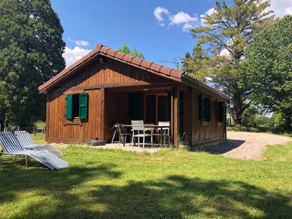 A wooden chalet is set within a green landscape, featuring a covered terrace with outdoor seating. Sun loungers are placed on the grass, and trees provide natural shade, contributing to a peaceful outdoor environment.