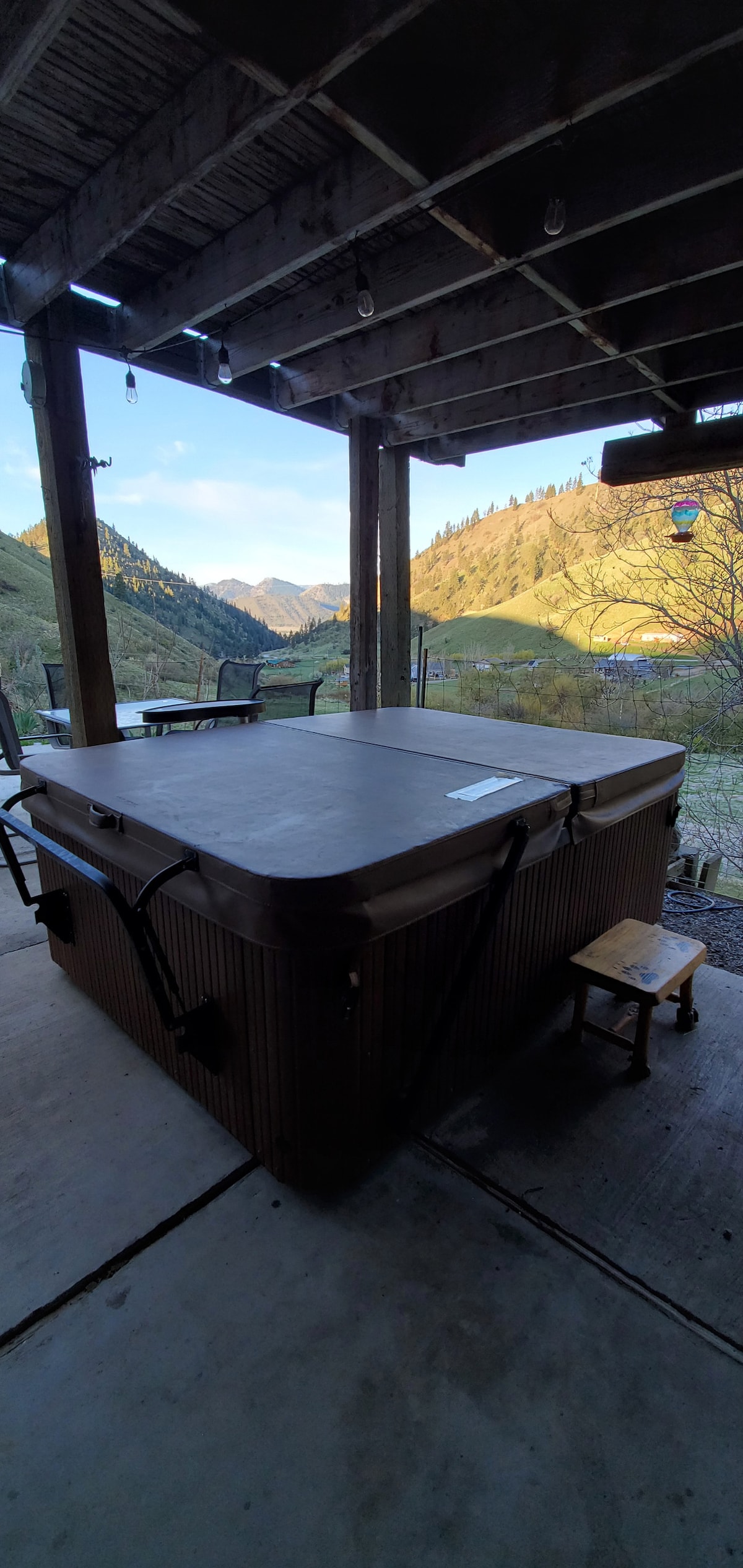 A hot tub sits on a covered patio, surrounded by mountain views. The structure's wooden beams and concrete flooring provide a rustic feel, with outdoor seating visible in the background. Soft light illuminates the landscape, highlighting the rolling hills and natural scenery.