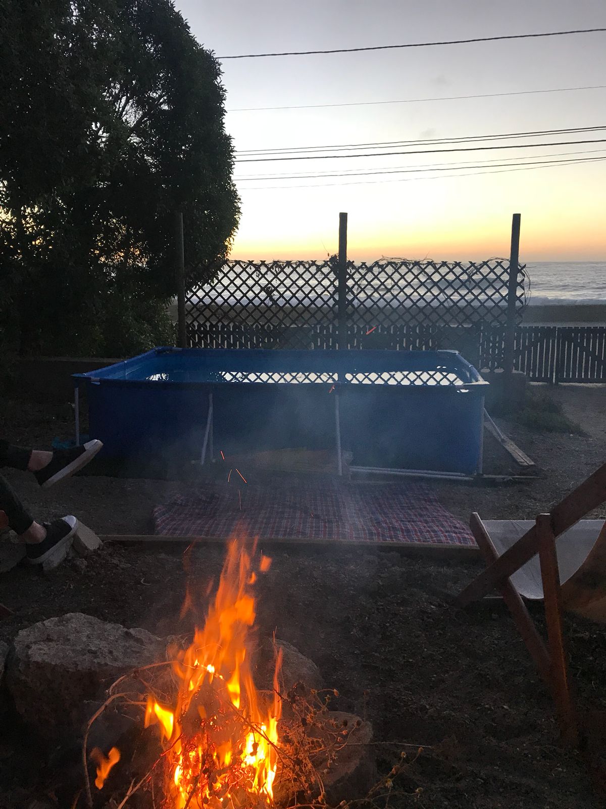 An outdoor space is shown with a small swimming pool and a fire pit. The fire burns steadily, casting gentle light. In the background, a sunset creates a soft gradient over the horizon, while a wooden fence encloses the area, ensuring privacy.