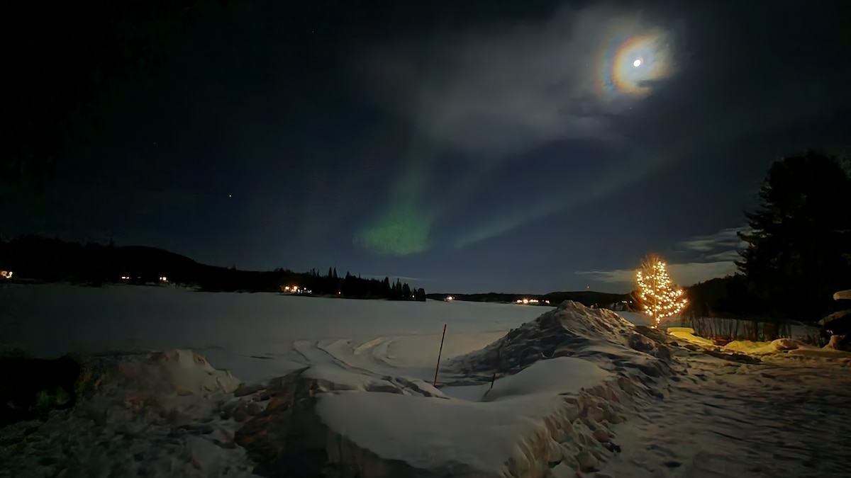 A serene winter landscape is shown under a night sky illuminated by the moon and an array of northern lights. Snow-covered ground features subtle topography, while a decorated tree is positioned nearby. Distant lights from nearby structures create a calm ambiance.
