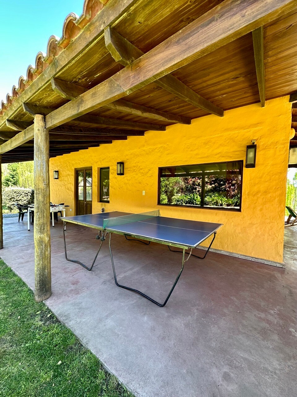 An outdoor area features a ping pong table under a shaded porch with wooden beams. The vibrant yellow walls of the adjacent structure contrast with the green grass, while seating arrangements are visible in the background, promoting a relaxed environment.