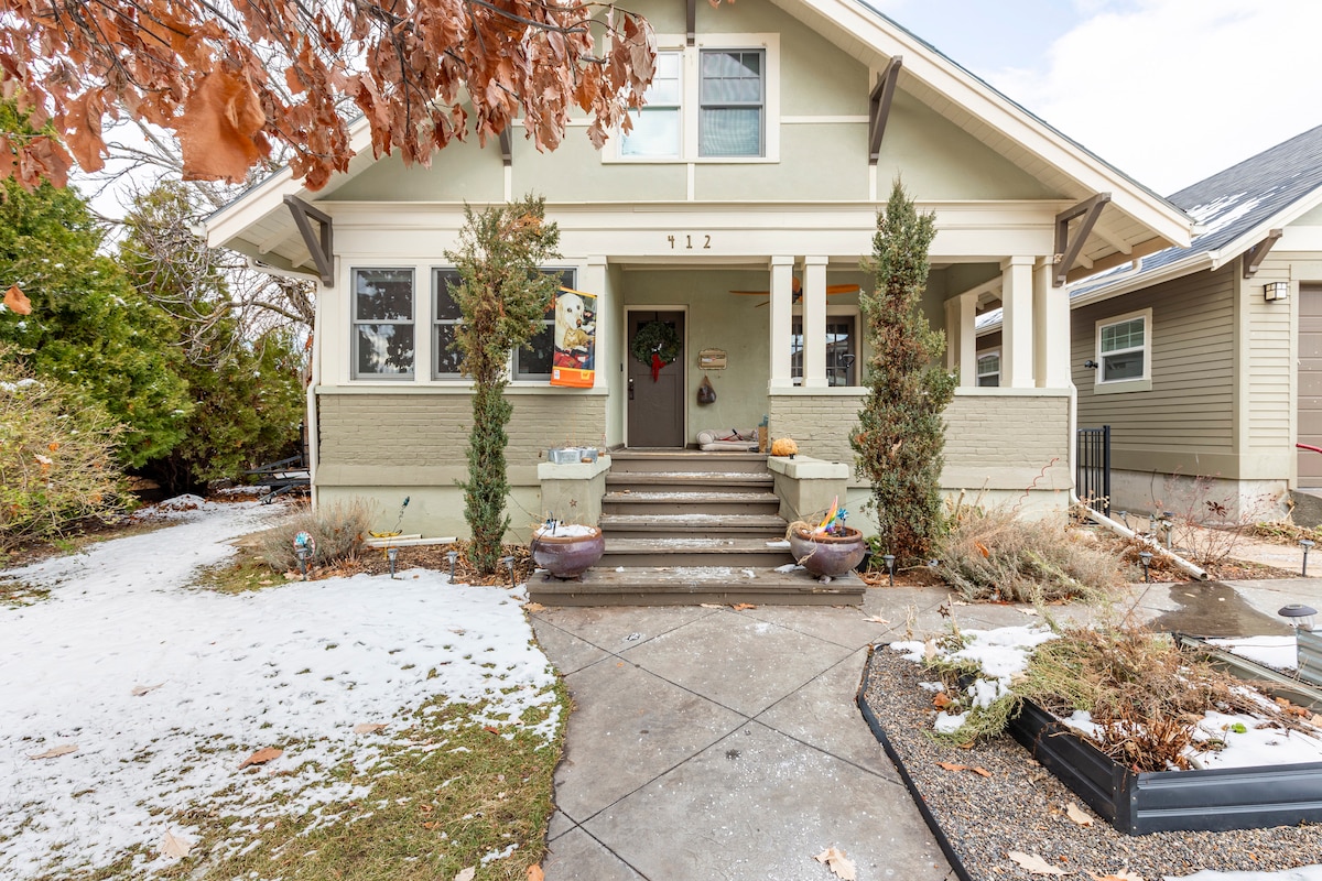 A spacious front porch features steps leading up to the entrance of a light green house. Flanking the porch are neatly trimmed shrubs and seasonal decorations. A light dusting of snow covers the ground, creating a calm winter scene.