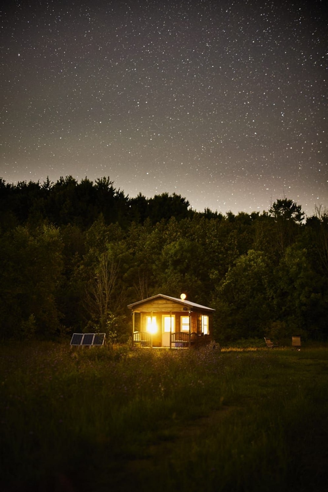 A cabin is illuminated by warm light against a starry night sky. Solar panels are visible nearby, with a backdrop of lush trees providing a sense of privacy. The grassy field surrounds the structure, emphasizing its secluded setting in nature.