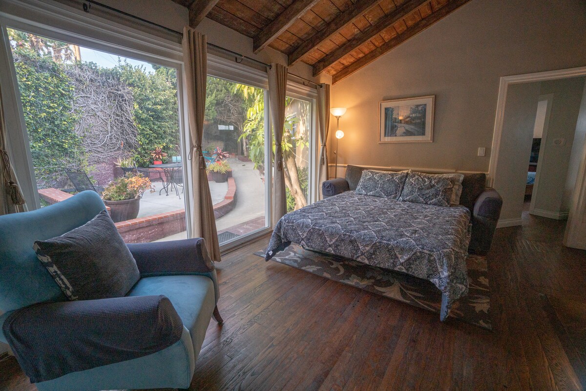 A spacious sleeping area features a comfortable bed with patterned bedding, situated next to a large window that invites natural light. A cozy chair is placed nearby, and the outdoor view includes greenery and a landscaped patio area.