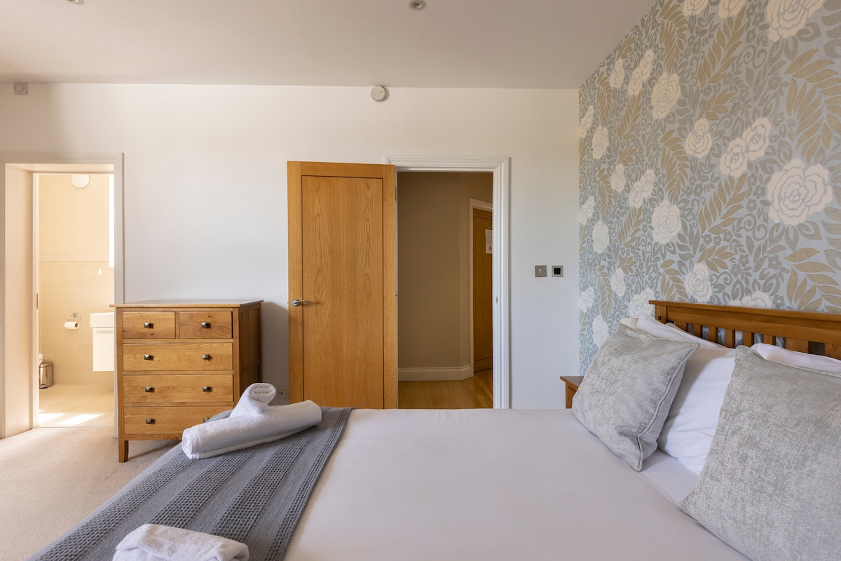 A spacious master bedroom is featured, showcasing a wooden bed with soft linens and decorative pillows. A light-colored floral accent wall complements the decor. A wooden dresser is positioned nearby, and natural light fills the room through the open door leading to the en-suite bathroom.