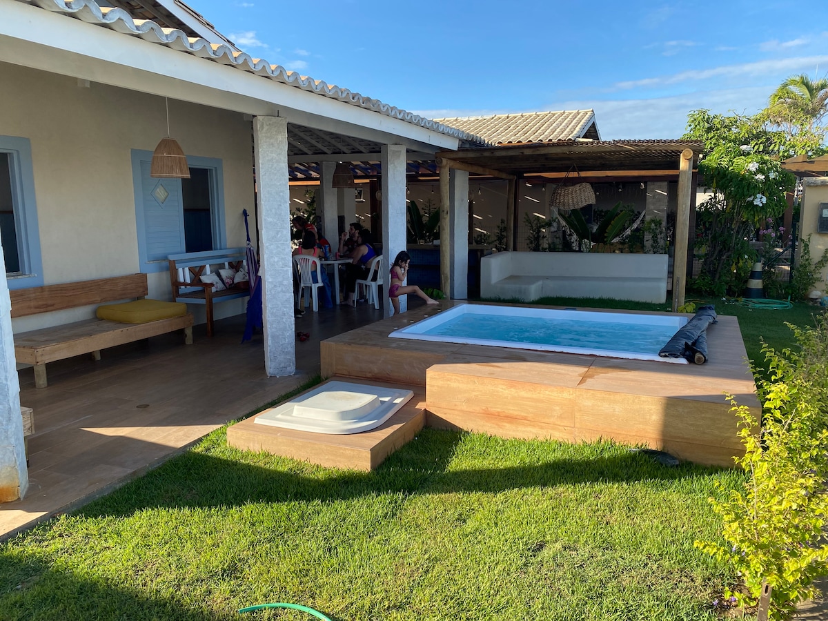 An outdoor area features a wooden deck with a spa tub, surrounded by lush greenery. The space includes a cozy seating area with cushions and a sheltered dining space, inviting relaxation and social gatherings. The bright sky complements the tranquil environment.