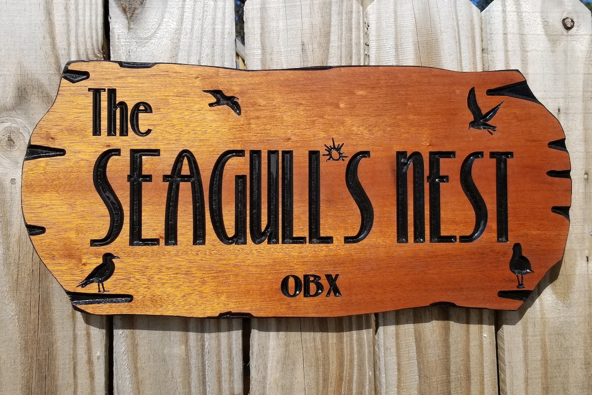 A wooden sign displaying the name 'The Seagull's Nest OBX' features carved lettering and silhouettes of birds. The sign is mounted on a weathered wooden fence, enhancing the rustic appeal of the surrounding area.