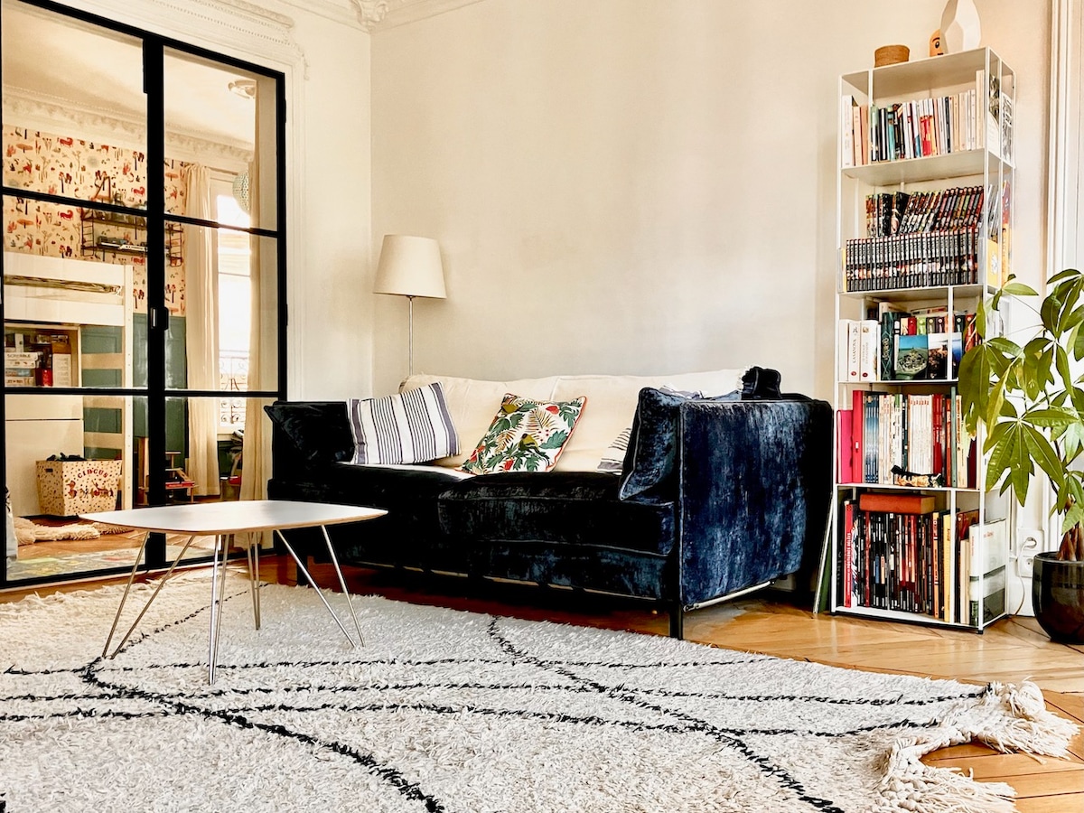 A living room is presented with a dark upholstered sofa featuring patterned cushions. A sleek coffee table is positioned in front of the sofa on a textured area rug. A tall bookshelf filled with books stands nearby, complemented by a decorative plant and soft light from a floor lamp.