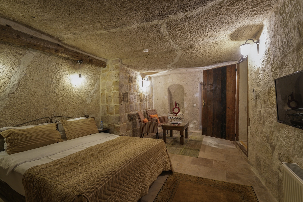 A cave suite is presented, featuring a spacious bed with textured bedding. Stone walls and a wooden beam ceiling provide a natural atmosphere. A small sitting area is seen with a wooden table and two chairs. Wall-mounted lighting enhances the cave's unique features.