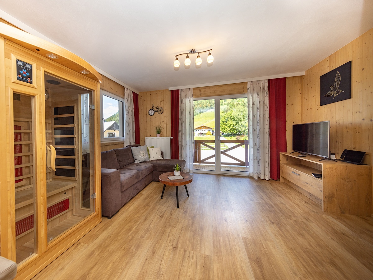 A bright living area features a comfortable couch and a coffee table, complemented by large sliding doors that open to a scenic balcony. A sauna is present in one corner, while a TV is mounted on the wall opposite the seating area.