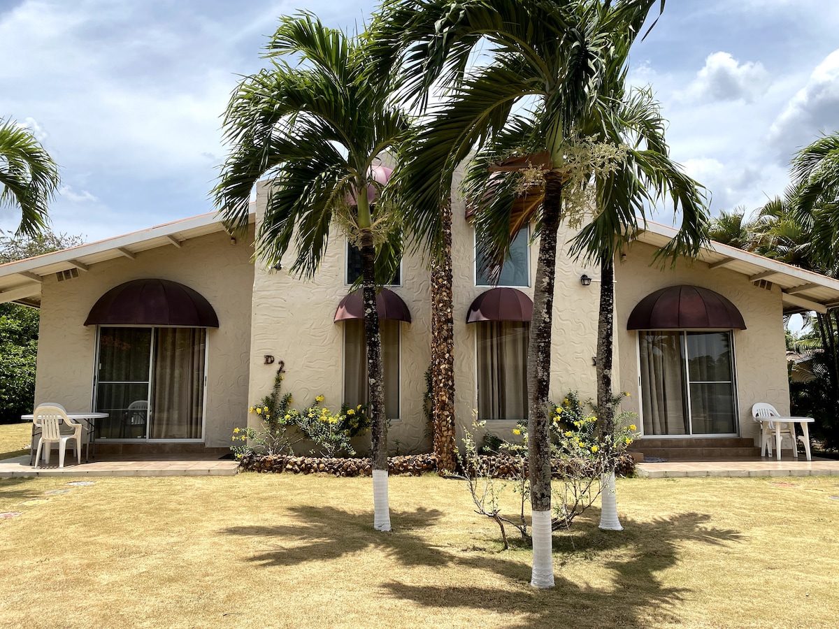 The exterior of a two-story building is presented, featuring distinctive brown awnings over large windows. A neat lawn is surrounded by palm trees, with two white chairs positioned in front of the entrances. Light clouds drift across a bright sky.