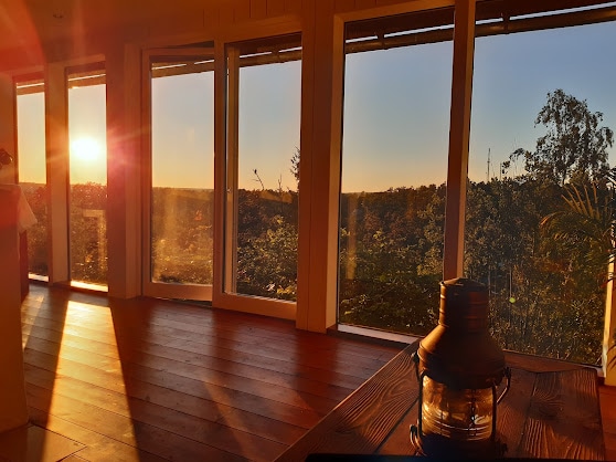 Sunlight streams through large glass windows, illuminating a spacious room with warm wooden flooring. A rustic lantern sits on a wooden table, creating a calming focal point, while a stunning view of greenery is framed by the glowing sunset.