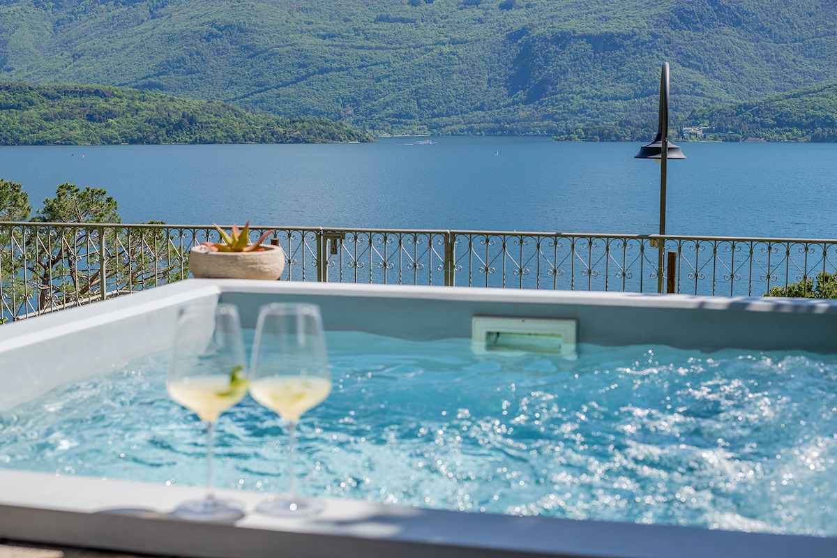 A hot tub is positioned on a patio overlooking the tranquil waters of the lake, with scenic mountains in the background. Two glasses filled with a clear beverage sit alongside the tub, inviting relaxation and enjoyment of the striking view.