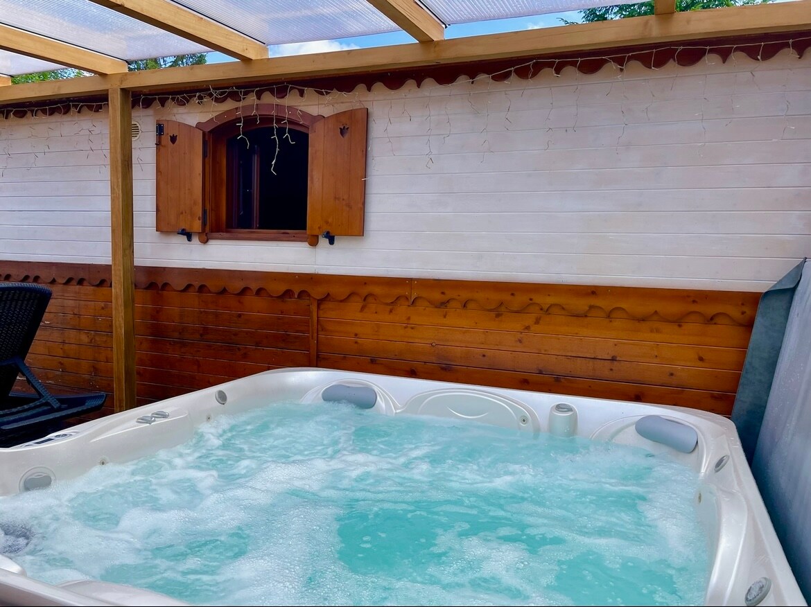A private jacuzzi is nestled under a wooden canopy, featuring bubbling water that creates a relaxing atmosphere. Surrounding wooden panels provide a cozy backdrop, while a window with wooden shutters allows for natural light. Subtle fairy lights add a soft glow to the space.