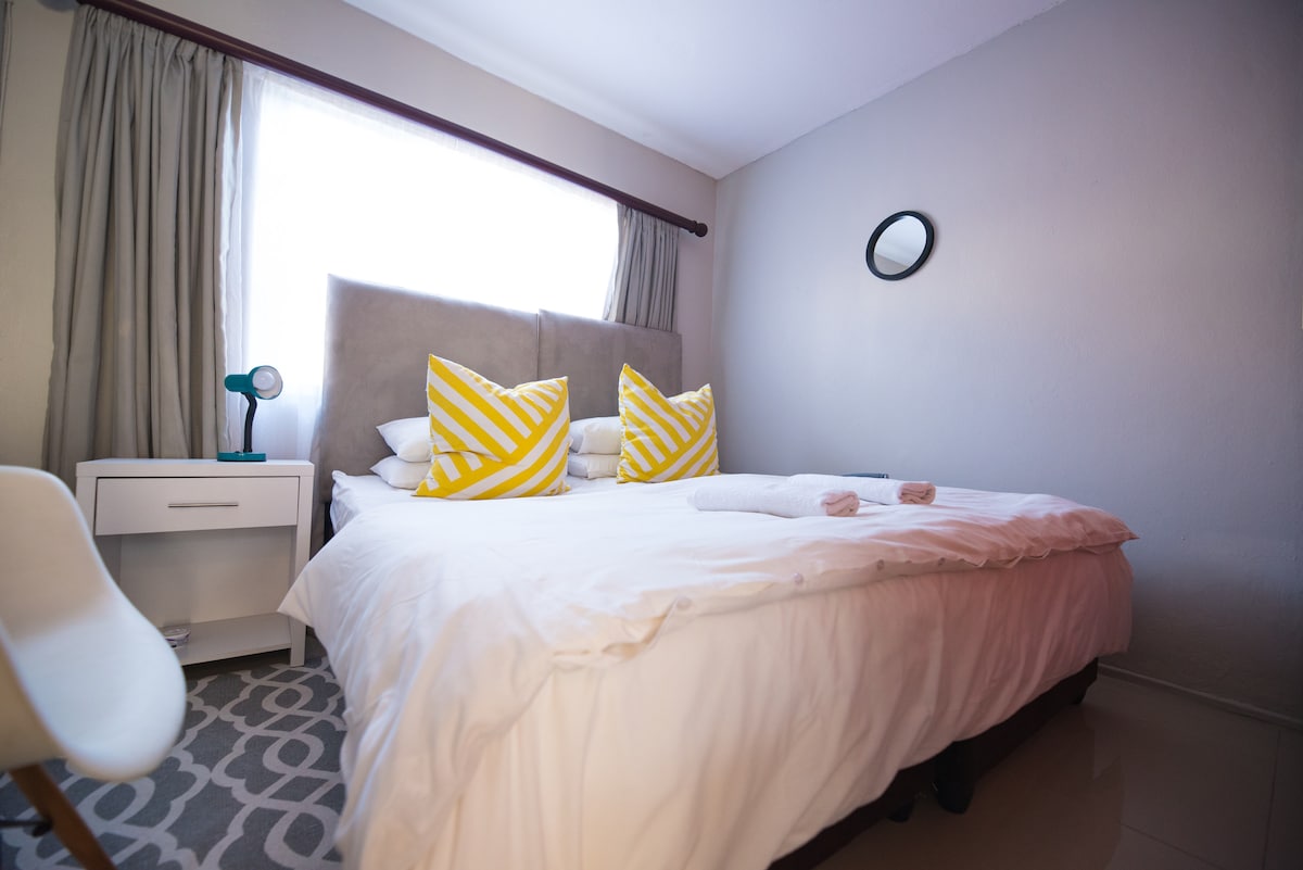 The bedroom features a spacious bed with soft white linens and two vibrant yellow accent pillows. Natural light fills the room through a large window, complemented by light gray curtains. A small bedside table and a simple chair add to the minimalist decor.