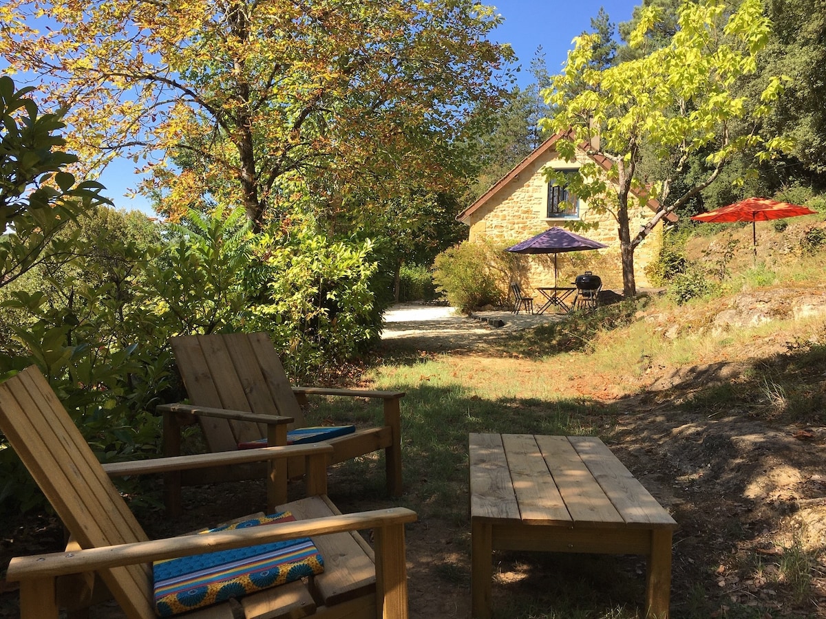 A garden area features two wooden chairs and a wooden table, surrounded by vibrant greenery and trees. The stone gîte is visible in the background, with outdoor seating under umbrellas of various colors, creating a serene setting for relaxation.