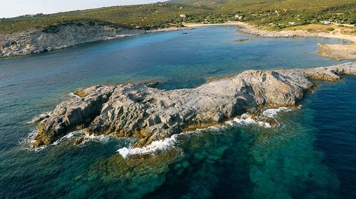 An aerial view of a coastal area reveals rocky formations jutting out into clear blue waters. The shoreline is bordered by lush green hills and features a sandy beach in the distance, complementing the natural landscape.