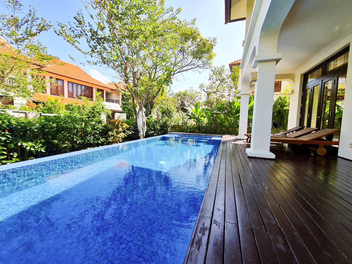 A serene private pool features calm blue waters, surrounded by lush greenery. A wooden deck provides space for relaxation, with two sun loungers positioned nearby. The scene is framed by vibrant trees and plants, creating a tranquil outdoor environment.