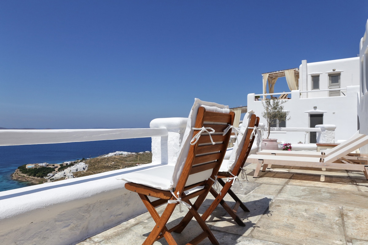 A spacious terrace features wooden loungers with soft cushions, offering views of the Aegean Sea and surrounding landscape. The bright blue sky complements the white architecture, creating a serene space for relaxation. Potted flowers add a touch of color to the inviting outdoor area.