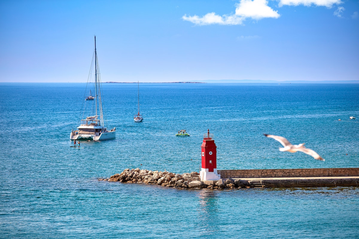 A serene coastal scene is depicted, featuring a bright red lighthouse standing on a stone pier. Various boats are seen in the calm blue waters, with a cloudy sky overhead. A seagull is captured in flight near the lighthouse, enhancing the maritime atmosphere.