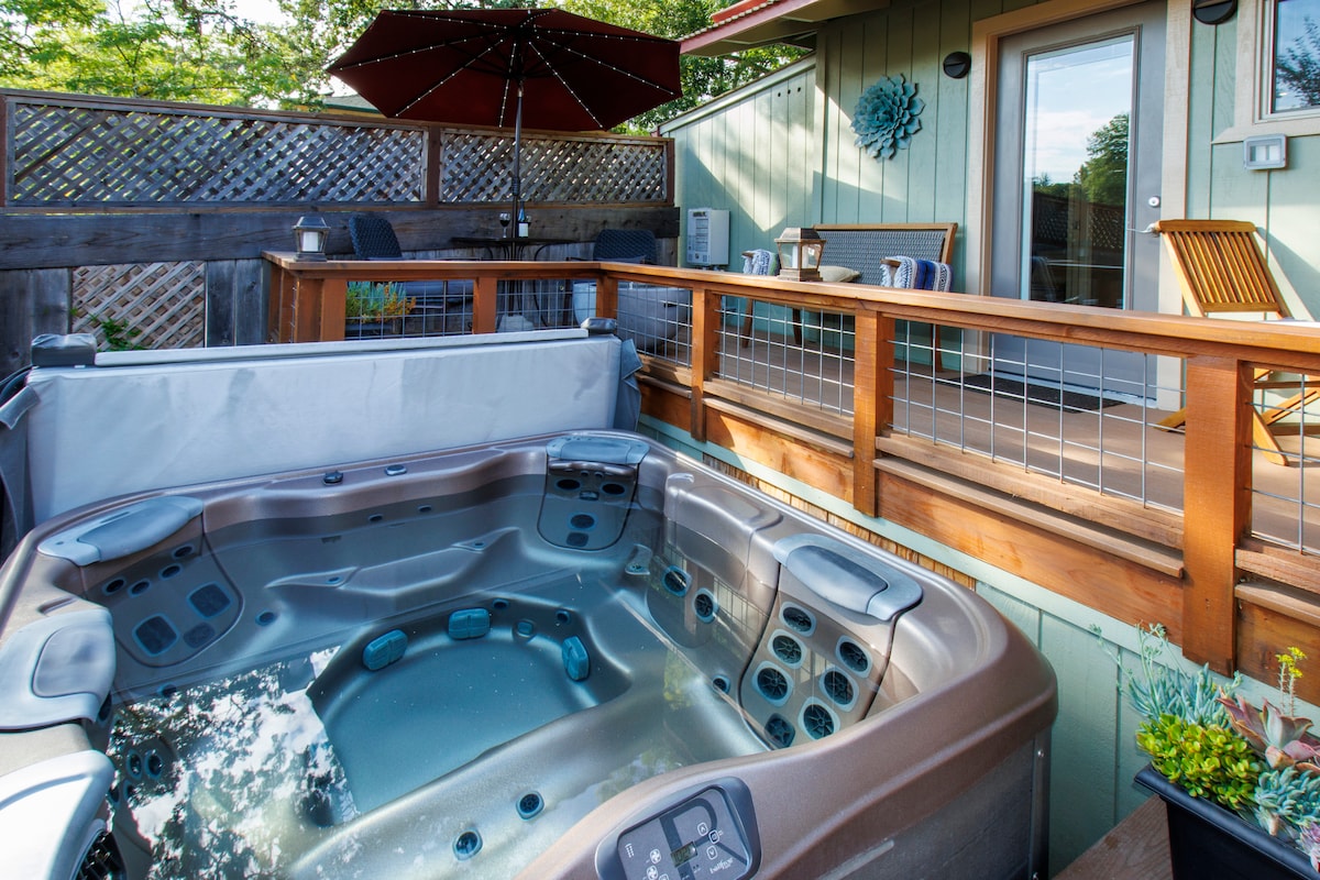 A spacious hot tub is situated on a private deck, surrounded by wooden railings and potted plants. A table with four chairs and a patio umbrella are visible in the background, leading to a glass door that opens to the guest suite.