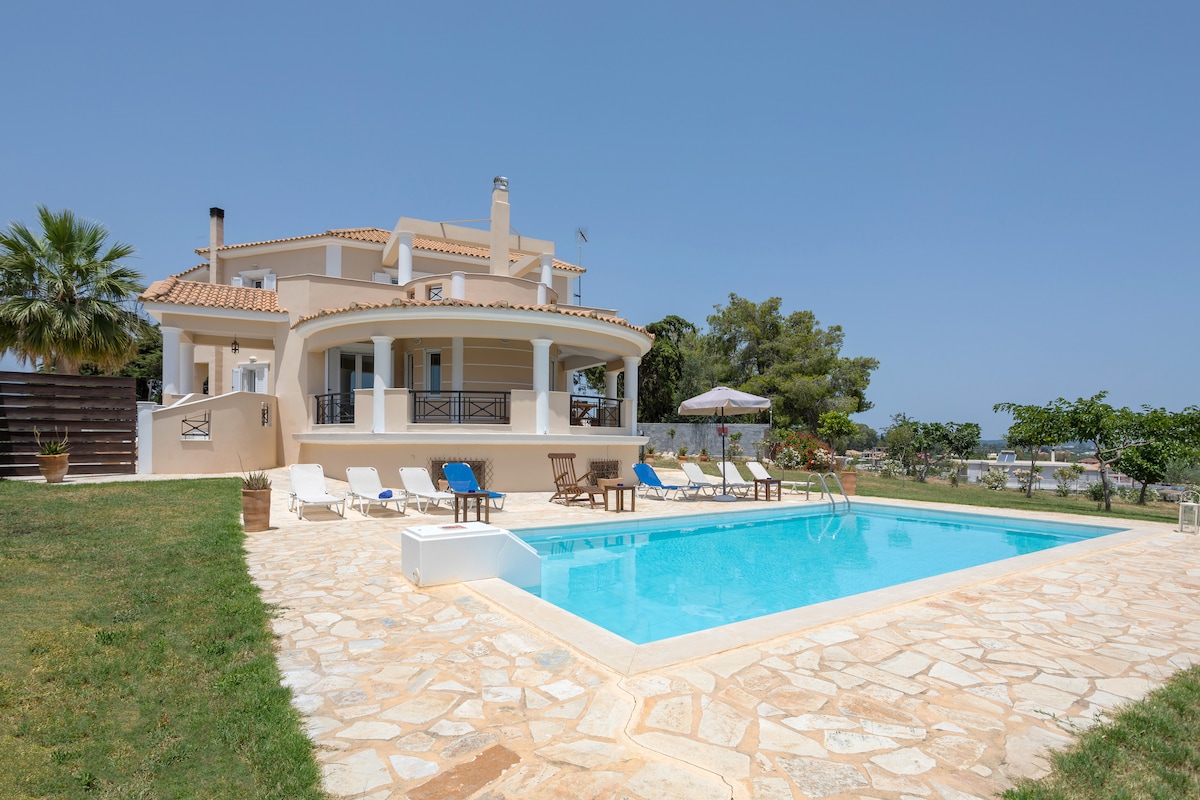The exterior of the villa showcases a private swimming pool surrounded by a stone patio. Comfortable sun-loungers are arranged around the pool, and a shaded seating area is visible. Lush greenery complements the setting, while the villa's two-story structure offers a welcoming presence.