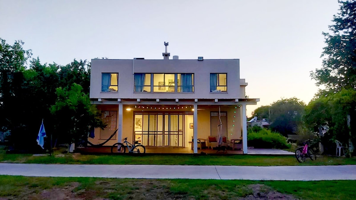 The exterior of a modern two-story house is shown, featuring large windows that allow soft evening light to fill the interior. Surrounding greenery adds a serene backdrop, with bicycles resting against the walls and a comfortable seating area visible on the porch.