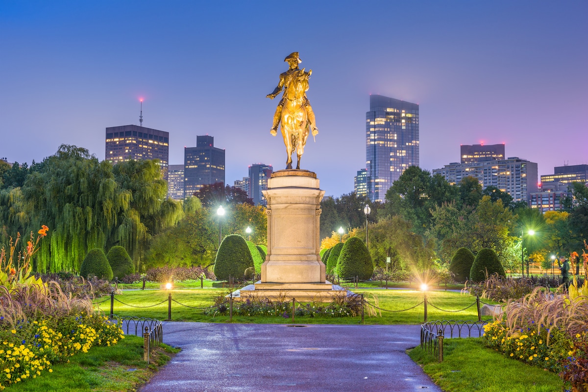 A statue of George Washington mounted on a horse stands prominently in a park, surrounded by neatly trimmed hedges and flower beds. The city skyline glows softly in the background, with lights twinkling against the evening sky.