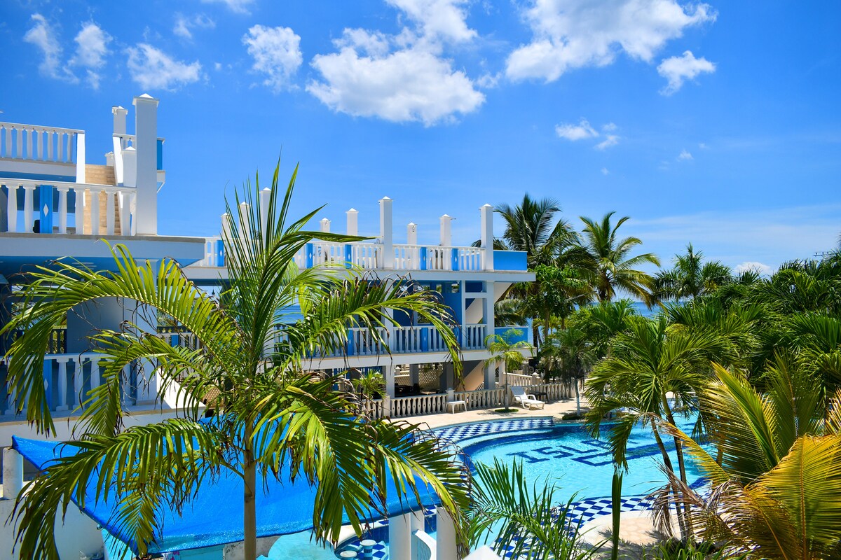 The property features a vibrant blue exterior with multiple levels, surrounded by lush palm trees. A spacious pool with a checkerboard pattern is visible, complemented by bright blue skies and fluffy white clouds. The design encourages a relaxed atmosphere in a tropical setting.