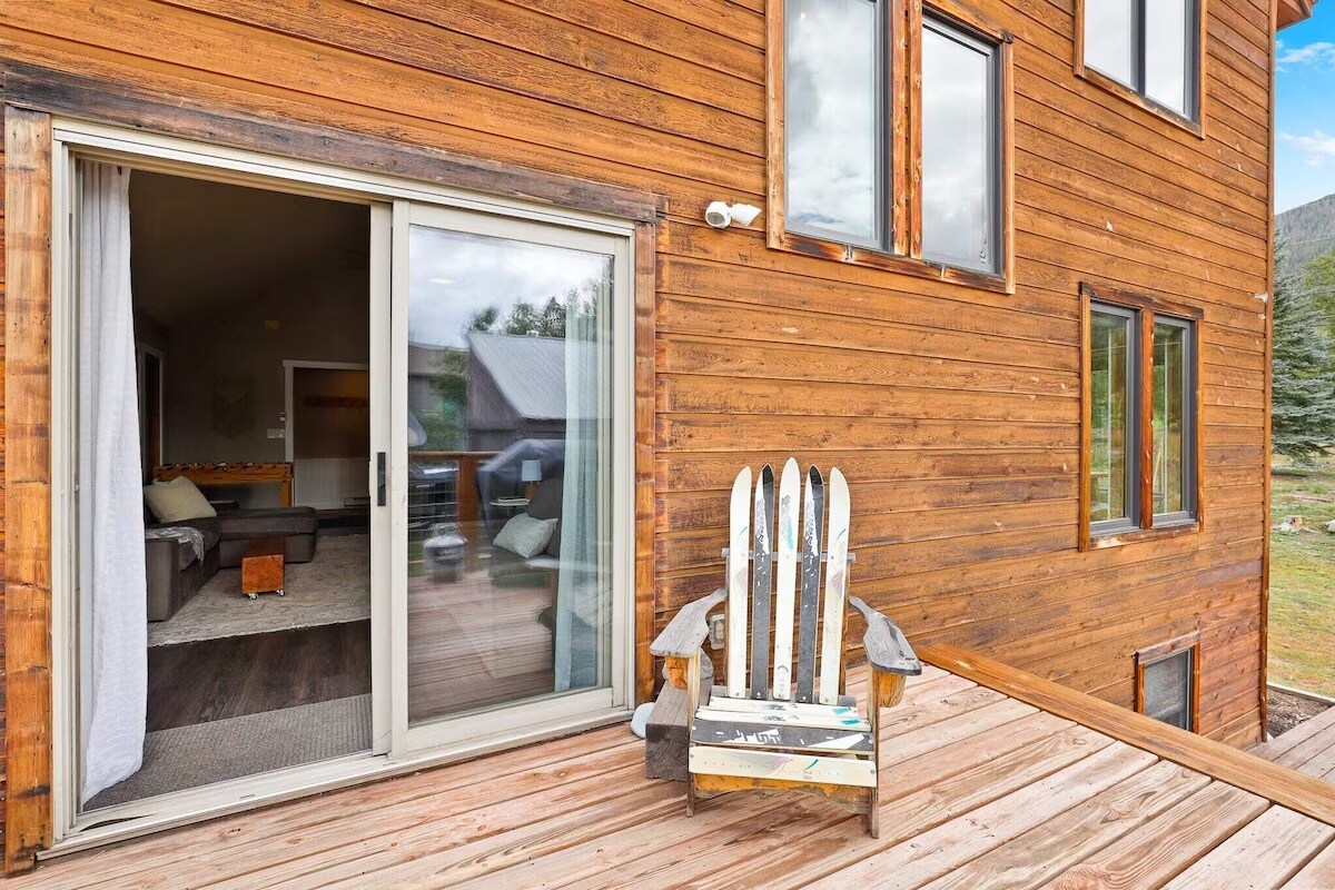 A wooden deck extends from the house, featuring a rustic chair designed for relaxation. Large sliding glass doors open to the indoor living space, offering glimpses of natural light and comfortable seating. A backdrop of mountain scenery complements the wooden exterior.
