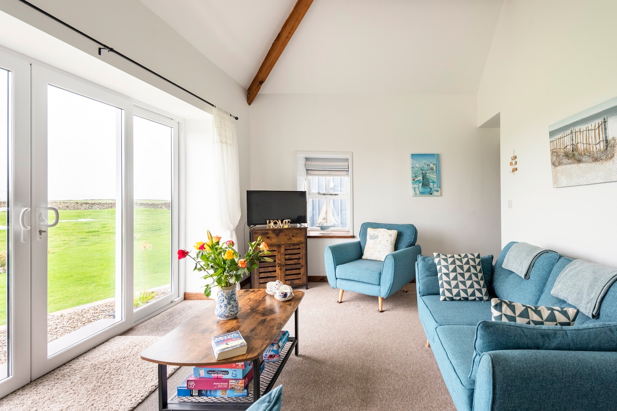 A spacious living area features large patio doors that provide views of the surrounding farmland. Comfortable seating is arranged around a wooden coffee table, adorned with a floral arrangement. A flat-screen TV and decorative artwork complete the room, highlighting a calm and inviting space.