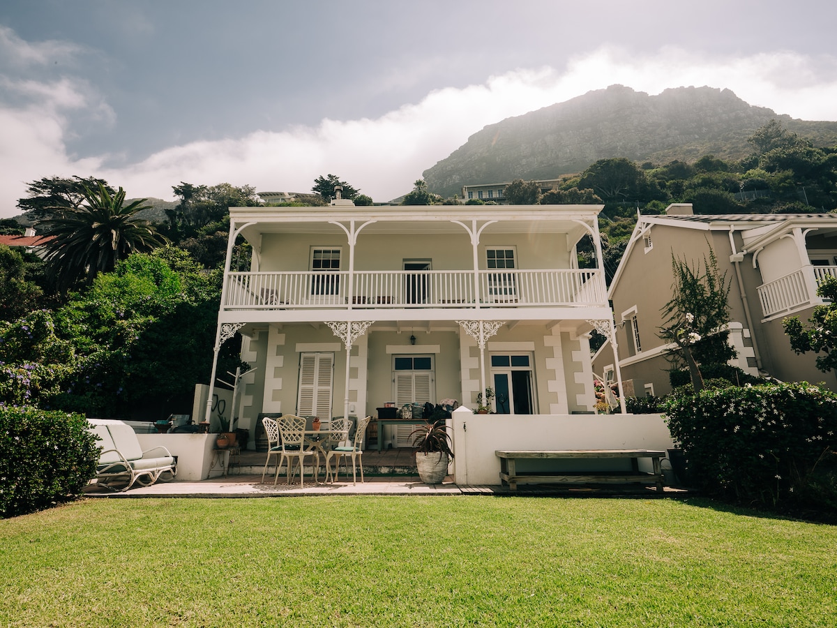 The Family Villa stands elegantly amidst lush greenery, featuring a lush, level garden in front. A spacious patio is furnished with outdoor seating, and a balcony extends from the upper level. The backdrop of a mountain enhances the serene coastal environment.