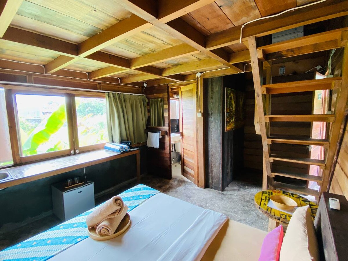 A spacious room is designed with wooden elements, featuring a comfortable bed and a small seating area. Large windows provide natural light and views of the green surroundings. A staircase leads to an upper level, while a small kitchenette can be seen in the corner.