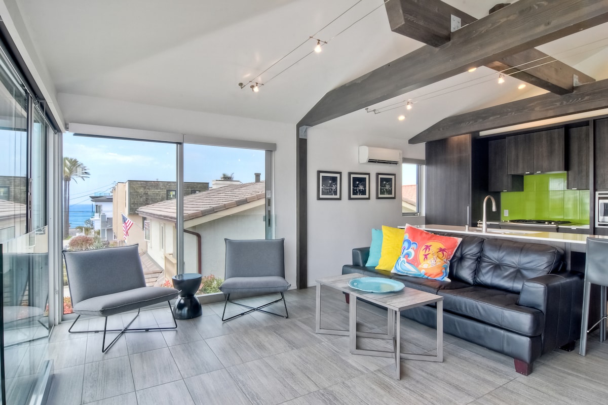 A modern living area is presented with large windows, offering views of the beach outside. Stylish seating includes two gray chairs and a black leather sofa adorned with colorful throw pillows. Natural light illuminates the space, highlighting the open floor plan and contemporary design.