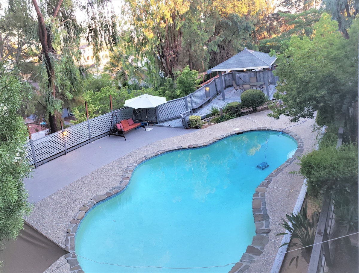 A private pool is surrounded by stone and concrete with a spacious deck. Greenery, including mature trees and shrubs, enhances the outdoor area. A lounge chair and an umbrella are positioned near the pool, providing a relaxing atmosphere for guests.