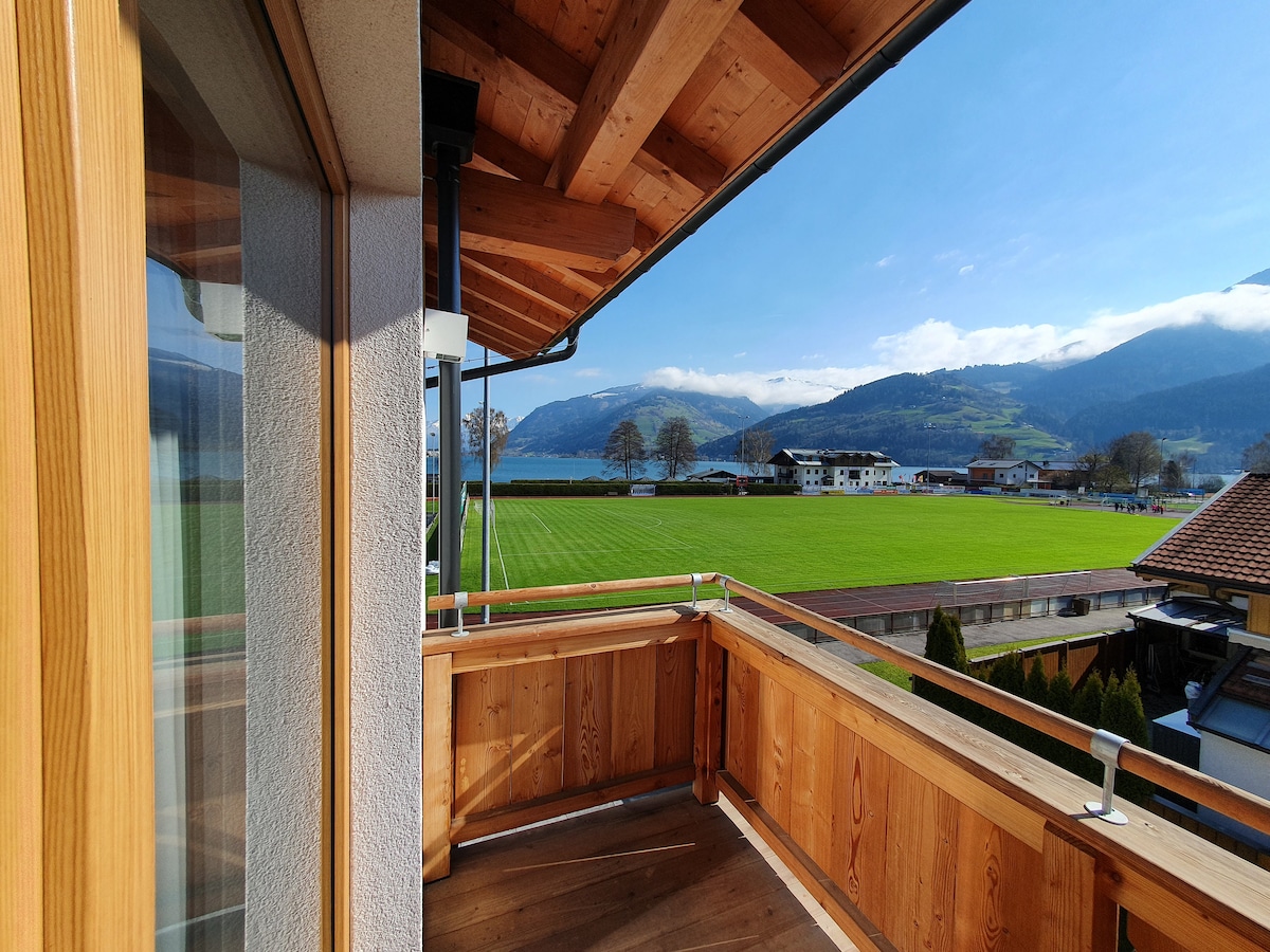A spacious wooden balcony features panoramic views of the surrounding landscape, including lush green fields and mountains in the distance. Natural light floods the area, highlighting the warm wood tones of the balcony's railing and floor.