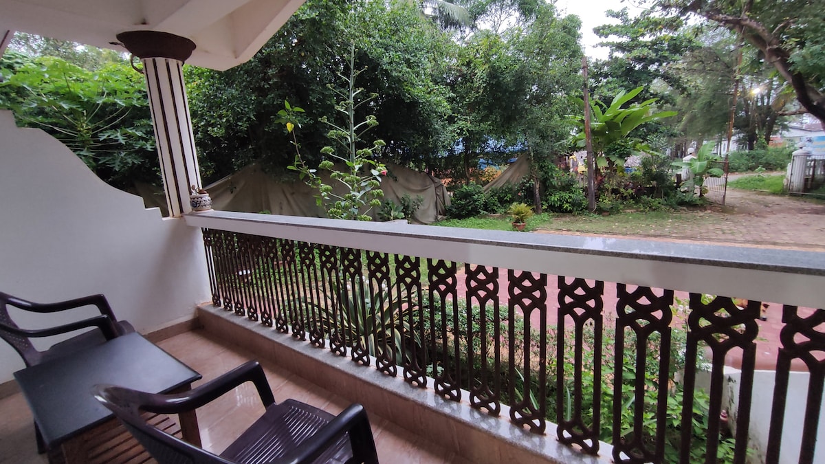 A balcony area features two black chairs overlooking a lush garden filled with greenery. The intricate railing design adds character, while the serene surroundings create a peaceful atmosphere. The scene is complemented by natural light filtering through the trees.