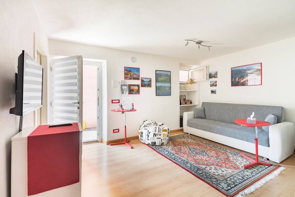 The living area features a light-colored wall with framed photographs of scenic views. A gray sofa and a patterned rug provide comfort. A small red table and a unique pouf add character. Natural light streams in through a partially open door leading outside.